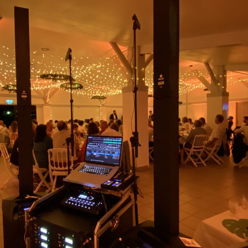 Des enceintes colonnes noires imposantes de chaque côté. Un poste de DJ avec un ordinateur portable affichant un logiciel de mixage et une table de mixage. Une salle de réception remplie d'invités, décorée de guirlandes lumineuses au plafond.