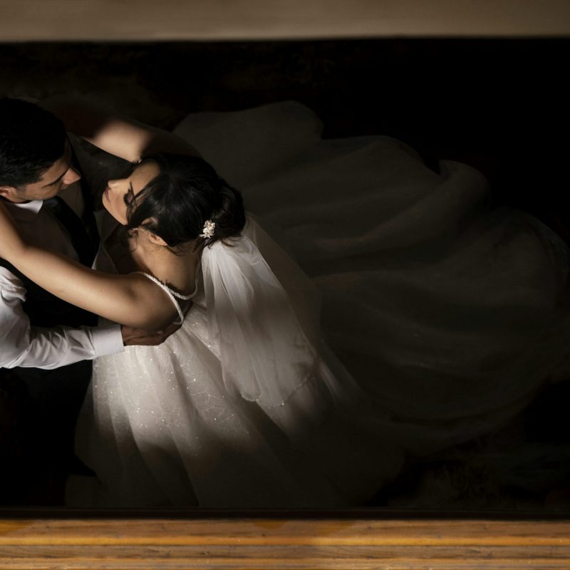 Vue de dessus d'un couple de mariés dansant dans un halo de lumière entouré d'obscurité. La mariée, dans une élégante robe blanche à voile, entoure le cou du marié de ses bras, tandis qu'ils se regardent intensément. La composition en clair-obscur accentue l'aspect romantique et intime de la scène.