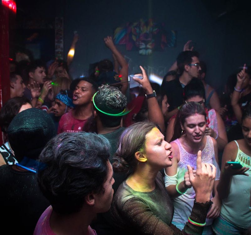 Une foule de jeunes gens danse et fait la fête dans une salle faiblement éclairée. Plusieurs personnes ont le visage ou les vêtements couverts de poudre colorée et de peinture fluorescente. L'ambiance est dynamique, avec des bras levés, des gens qui discutent et une personne qui regarde son téléphone au premier plan.