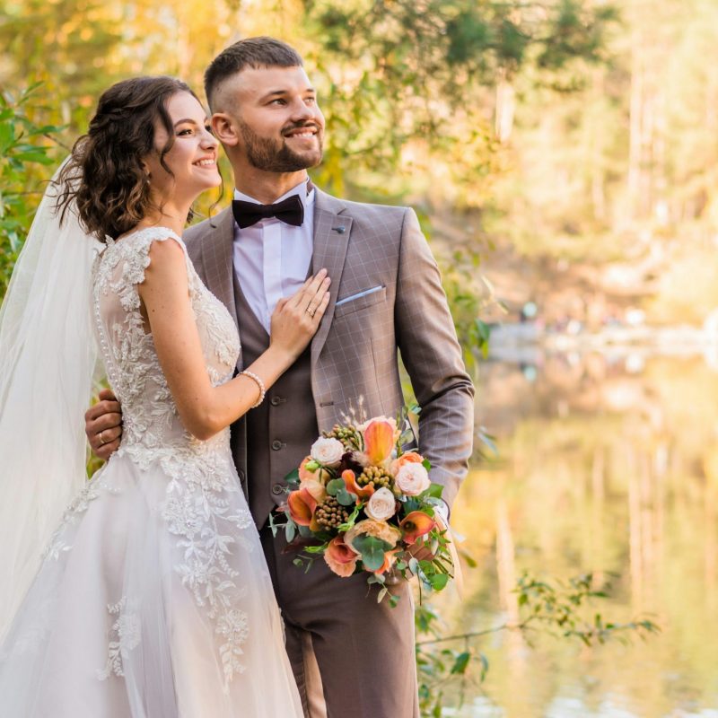 Un couple de jeunes mariés pose joyeusement au bord d'un lac entouré d'une forêt aux couleurs d'automne. La mariée porte une robe en dentelle blanche avec un bouquet de fleurs aux tons orange et crème, tandis que le marié porte un costume trois-pièces marron à carreaux avec un nœud papillon.