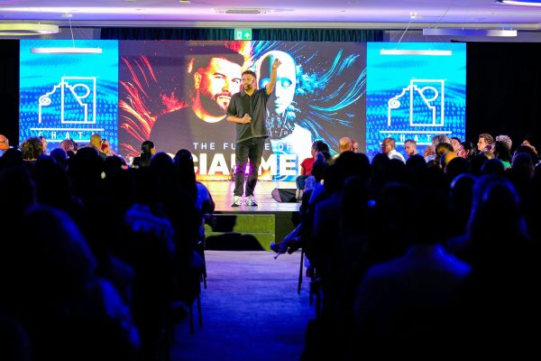 Un homme donne une conférence sur scène devant une large audience plongée dans l'obscurité. Derrière lui, un grand écran affiche un visuel futuriste représentant un visage humain et un robot, avec le texte « THE FUTURE OF SOCIAL MEDIA ». L'orateur a un bras levé, captivant l'attention du public dans une ambiance technologique et professionnelle.