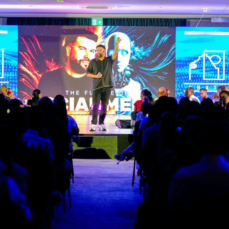 Un homme donne une conférence sur scène devant une large audience plongée dans l'obscurité. Derrière lui, un grand écran affiche un visuel futuriste représentant un visage humain et un robot, avec le texte « THE FUTURE OF SOCIAL MEDIA ». L'orateur a un bras levé, captivant l'attention du public dans une ambiance technologique et professionnelle.