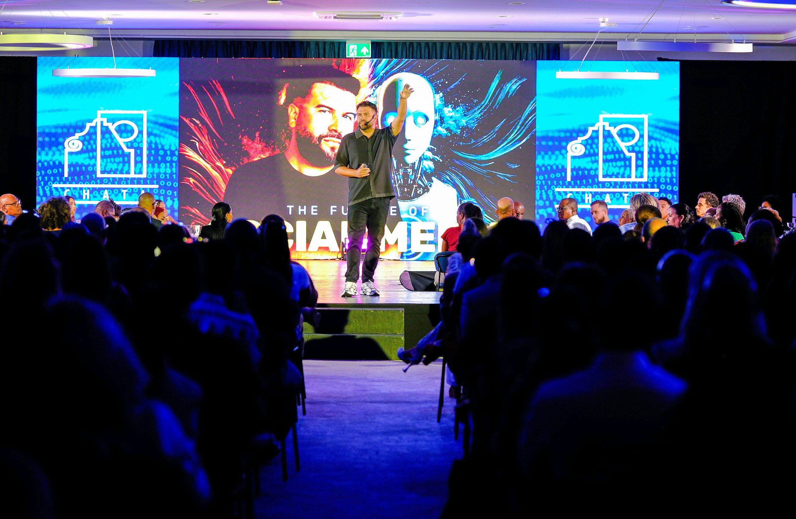 Un homme donne une conférence sur scène devant une large audience plongée dans l'obscurité. Derrière lui, un grand écran affiche un visuel futuriste représentant un visage humain et un robot, avec le texte « THE FUTURE OF SOCIAL MEDIA ». L'orateur a un bras levé, captivant l'attention du public dans une ambiance technologique et professionnelle.