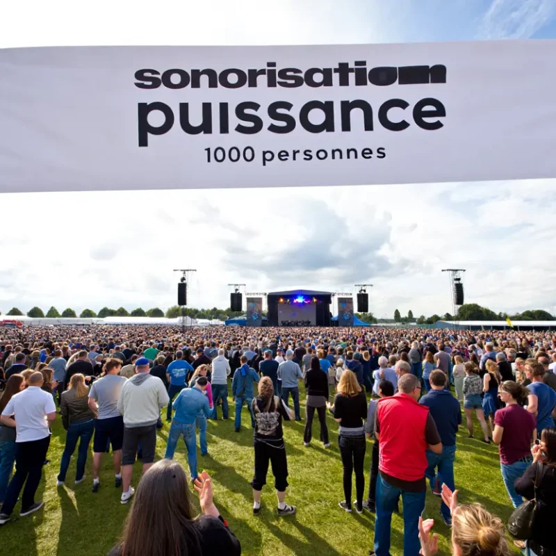 Une foule immense rassemblée dans un champ pour un concert en plein air, avec une grande banderole au premier plan indiquant "sonorisation puissance 1000 personnes".
