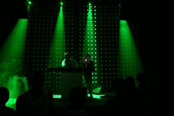 Deux DJs se produisent sur une scène plongée dans l'obscurité, illuminée par des faisceaux de projecteurs vert intense et un mur de LED à l'arrière-plan. On aperçoit la silhouette du public au premier plan, créant une ambiance de concert ou de club électro dynamique et immersive.