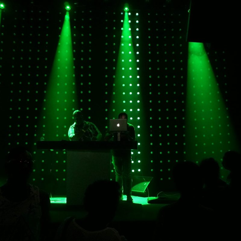 Deux DJs se produisent sur une scène plongée dans l'obscurité, illuminée par des faisceaux de projecteurs vert intense et un mur de LED à l'arrière-plan. On aperçoit la silhouette du public au premier plan, créant une ambiance de concert ou de club électro dynamique et immersive.