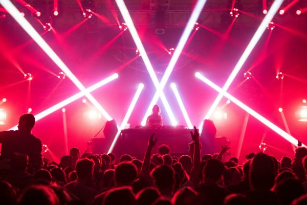 DJ en sets sur scène avec projecteurs laser blancs traversant la salle rouge, public en fête devant la scène.