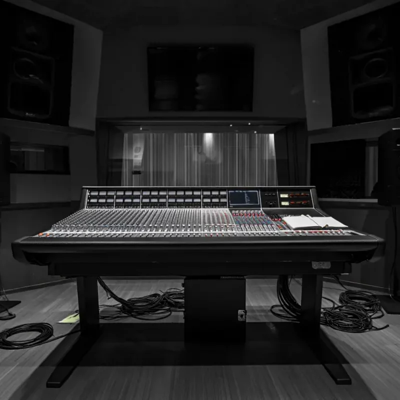 Une vue symétrique d'un studio d'enregistrement professionnel en noir et blanc. Au centre, une large console de mixage analogique trône face à une vitre donnant sur une cabine de prise de son. De chaque côté, deux enceintes de monitoring sur pieds encadrent l'installation. Le sol est jonché de câbles audio enroulés, renforçant l'aspect technique et créatif du lieu.