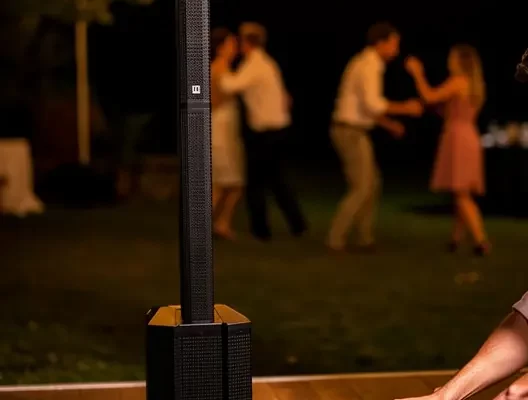 Paire de haut-parleurs de sono en extérieur avec DJ mixant sur une table, guirlandes lumineuses et personnes qui dansent en arrière-plan.