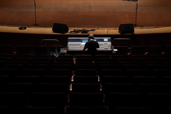 Vue en plongée d'un ingénieur du son assis devant une grande console de mixage numérique dans une salle de spectacle vide et sombre. L'ingénieur, vu de dos et portant des écouteurs, est concentré sur les nombreux faders, boutons et écrans lumineux de la console, tandis que des rangées de sièges vides s'étendent devant lui jusqu'à la scène, en partie éclairée, où sont installés des haut-parleurs et des supports d'éclairage. L'atmosphère est calme et studieuse, avec des lumières chaudes et des ombres douces.