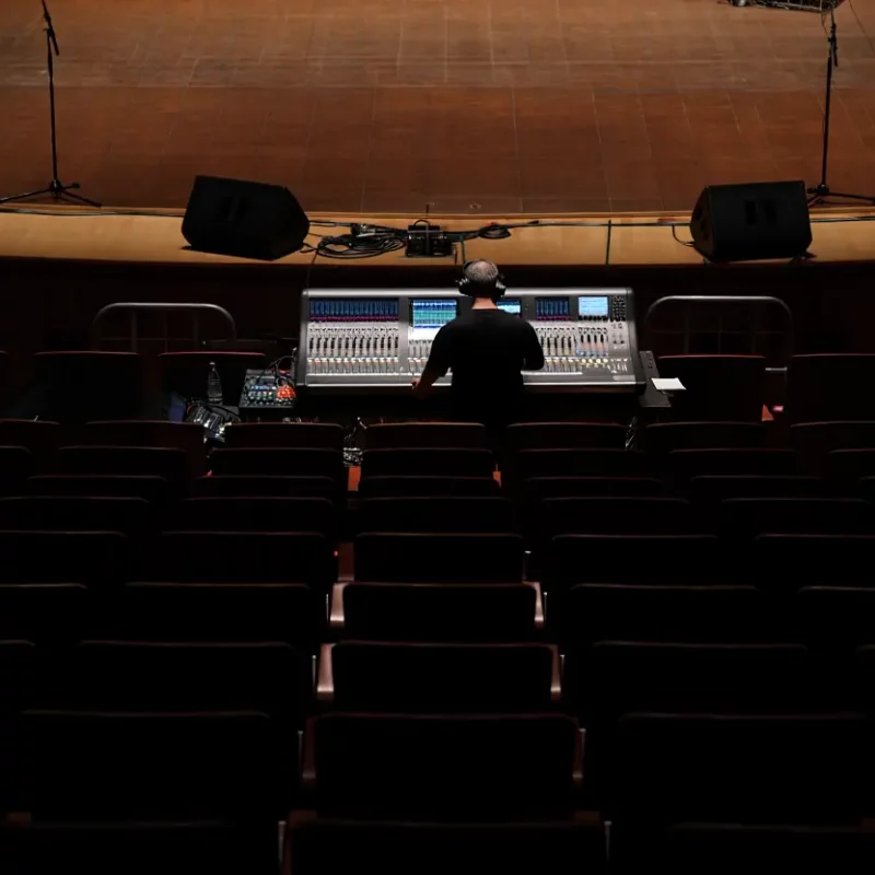 Vue en plongée d'un ingénieur du son assis devant une grande console de mixage numérique dans une salle de spectacle vide et sombre. L'ingénieur, vu de dos et portant des écouteurs, est concentré sur les nombreux faders, boutons et écrans lumineux de la console, tandis que des rangées de sièges vides s'étendent devant lui jusqu'à la scène, en partie éclairée, où sont installés des haut-parleurs et des supports d'éclairage. L'atmosphère est calme et studieuse, avec des lumières chaudes et des ombres douces.