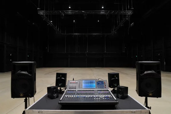 Une console de mixage numérique professionnelle avec écran tactile central, installée sur un flight case noir dans un grand studio de répétition ou une salle de spectacle vide. La console est entourée de deux paires d'enceintes de monitoring et de deux grandes enceintes sonorisées sur pieds. Au-dessus de la scène, une structure de pont technique en métal (truss) est suspendue au plafond noir.