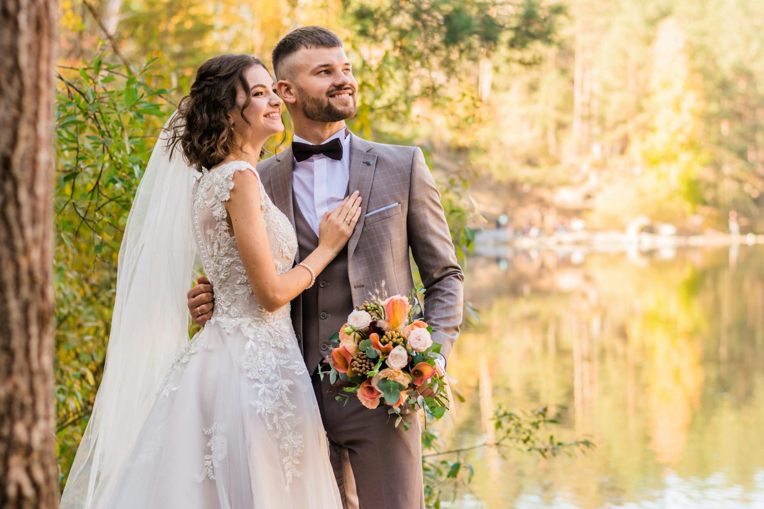 Mariée et marié souriants en extérieur près d’un lac, elle porte une robe blanche dentelle et voile; lui en costume gris et nœud papillon, bouquet floral à la main. Mariée et marié souriants en extérieur près d’un lac, elle porte une robe blanche dentelle et voile; lui en costume gris et nœud papillon, bouquet floral à la main.