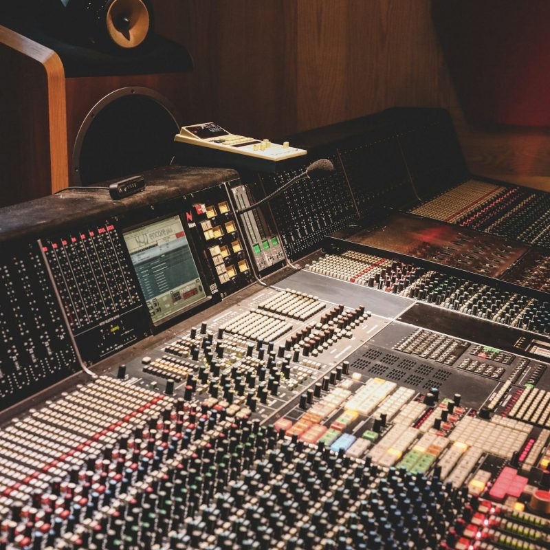 Une console de mixage analogique massive et détaillée remplit le cadre dans un studio d'enregistrement professionnel. Elle est dotée d'innombrables boutons, faders et touches lumineuses colorées. À l'arrière-plan, on aperçoit une enceinte de monitoring en bois de haute qualité dans un environnement aux boiseries chaleureuses.