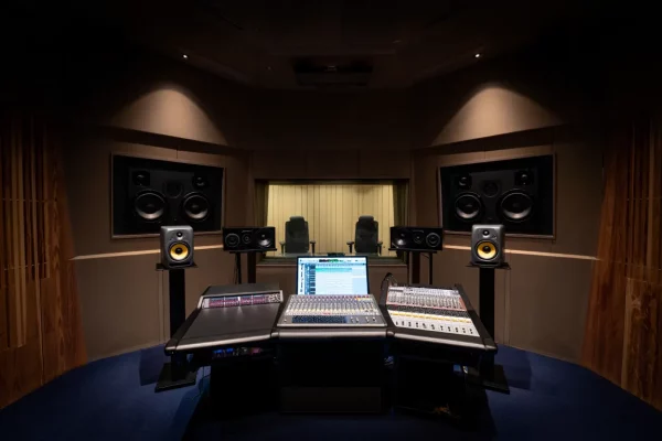 Une cabine de mixage de studio d'enregistrement haut de gamme à l'acoustique traitée. Au centre, une large console de mixage hybride avec un écran d'ordinateur affichant un logiciel de production audio. Plusieurs paires d'enceintes de monitoring sont disposées : de grandes enceintes intégrées aux murs et des enceintes plus petites (dont des KRK à membrane jaune) sur pieds. Au fond, une vitre donne sur une salle d'enregistrement où l'on aperçoit deux sièges vides.