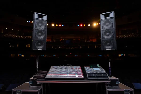 Une console de mixage audio et une platine DJ posées sur une table de régie, encadrées par deux colonnes d'enceintes imposantes dans une salle de spectacle plongée dans l'obscurité.