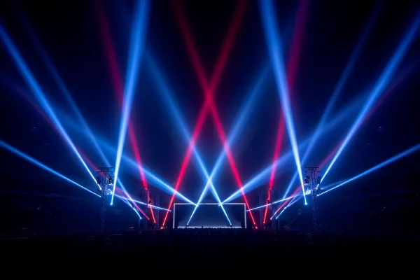 Des faisceaux lumineux bleus et rouges s'entrecroisent au-dessus d'une scène plongée dans le noir, créant un motif géométrique dynamique avec des jeux d'ombres et de lumière.