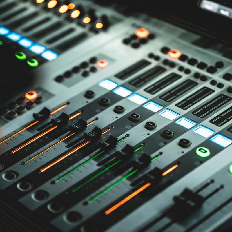 Gros plan sur une console de mixage numérique moderne avec plusieurs faders coulissants et des boutons rétroéclairés en bleu, vert et orange. La mise au point est nette sur les commandes centrales, tandis que le reste de la table de mixage s'estompe dans un flou artistique, créant une atmosphère technologique et créative.