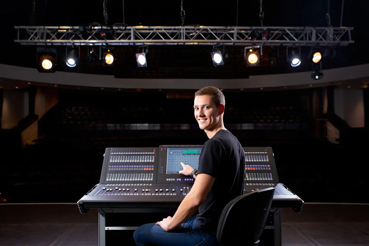Un technicien sourit en s'asseyant devant une console de mixage professionnelle dans un théâtre sombre, avec des projecteurs de scène allumés au-dessus de lui.