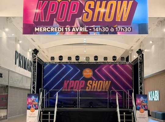 Une scène de spectacle compacte installée dans l'allée d'un centre commercial, prête pour un « KPOP SHOW ». Au-dessus de la scène est suspendue une immense bannière colorée affichant trois jeunes femmes au style K-pop et les informations de l'événement (mercredi 15 avril de 14h30 à 17h30). La scène elle-même est équipée d'un fond de scène assorti, de projecteurs et de garde-corps. En arrière-plan, on aperçoit les enseignes des magasins Primark et Kiabi.