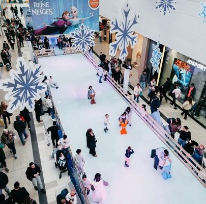 Une vue en plongée d'une patinoire éphémère installée dans l'allée centrale d'un centre commercial. Plusieurs enfants et adultes patinent sur la glace synthétique, certains utilisant des aides au patinage en forme d'animaux orange. La structure est décorée sur le thème du film Disney « La Reine des Neiges », avec de grands flocons de neige suspendus au plafond et une large bannière publicitaire représentant les personnages Anna et Elsa à l'extrémité de la piste. Des passants circulent de chaque côté de la patinoire devant les vitrines des magasins.