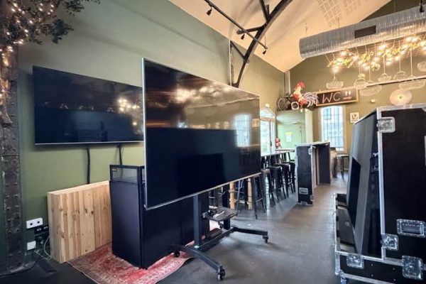 Une vue en perspective de l'intérieur d'un espace événementiel ou d'un bar moderne au style industriel. Au premier plan, un grand écran plat est monté sur un support mobile à roulettes. Derrière, un second écran est fixé sur un mur vert olive. Le décor comprend un faux arbre orné de guirlandes lumineuses, des poutres de toit noires apparentes, des flight-cases de transport et une enseigne lumineuse