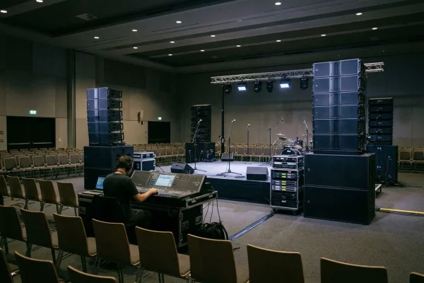 Vue d'ensemble d'une salle de conférence ou de spectacle en cours de préparation. Au premier plan, un technicien de dos est assis devant une console de mixage audio imposante. Face à lui, une scène éclairée est équipée d'une batterie, de micros sur pieds et d'une structure de projecteurs. De chaque côté de la scène, d'imposantes colonnes d'enceintes professionnelles (line array) sont empilées. La salle est remplie de rangées de chaises vides, créant une atmosphère de préparation technique avant un événement.