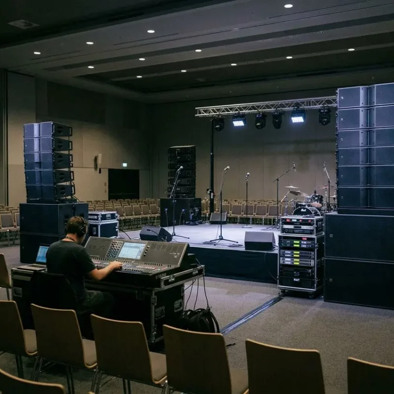 Une salle de conférence ou de spectacle spacieuse, montrant une installation de sonorisation professionnelle imposante. Au premier plan, un technicien de dos est assis devant une console de mixage numérique complexe. La scène, située à l'arrière-plan, est équipée de : Deux colonnes massives de haut-parleurs (systèmes line array) posées sur des caissons de basse imposants de chaque côté. Une structure de scène (truss) supportant des projecteurs d'éclairage. Plusieurs micros sur pieds et une batterie installée sur la plateforme. Des rangées de chaises vides qui font face à la scène, suggérant les préparatifs d'un événement de grande envergure.