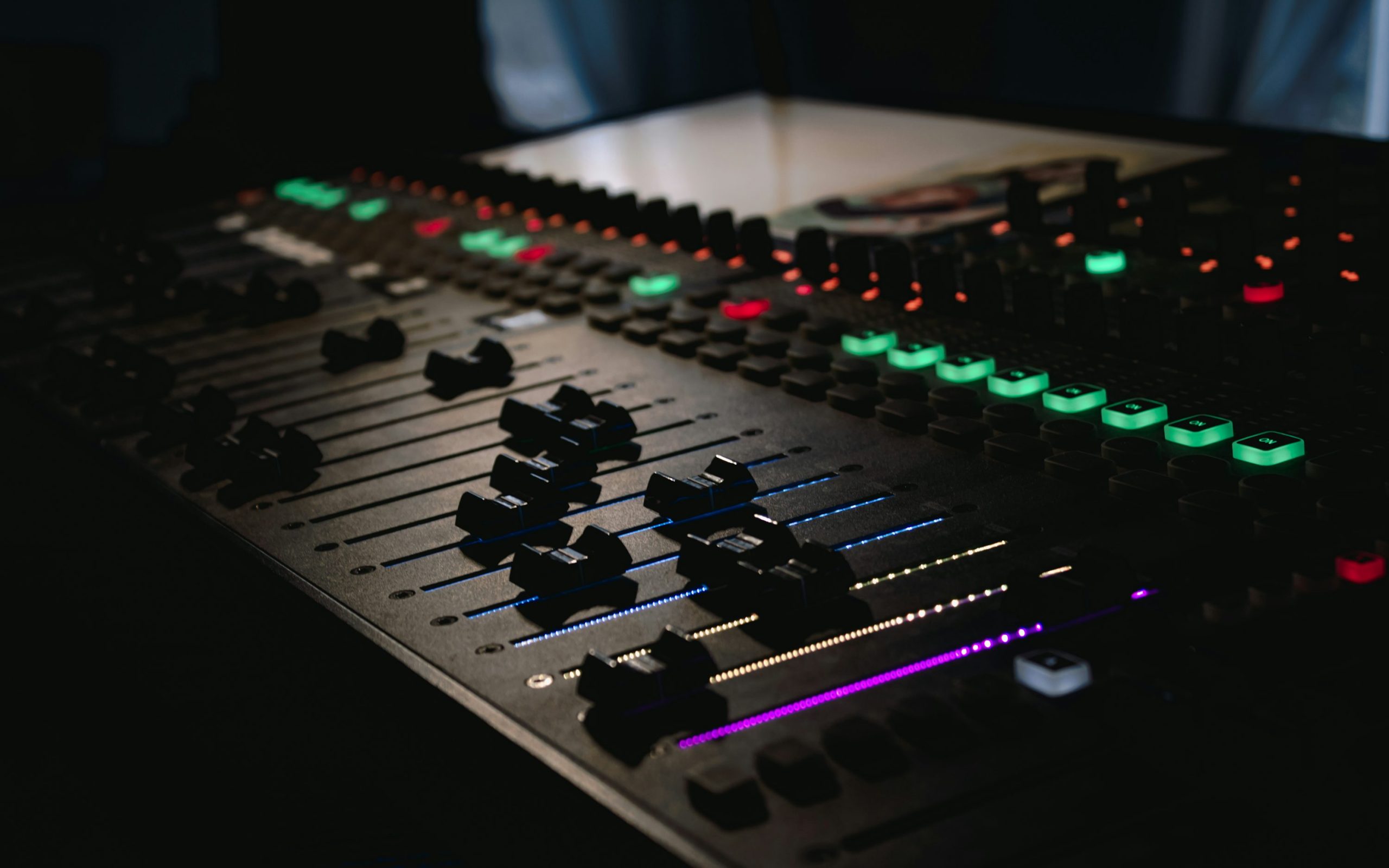 Table de mixage audio professionnelle avec voyants LED colorés et curseurs sombre, éclairage RGB sur console. (Main keyword: audio) Console de mixage audio-visuelle avec faders et boutons lumineux colorés, ambiance sombre. Location matériel audiovisuel Bordeaux.