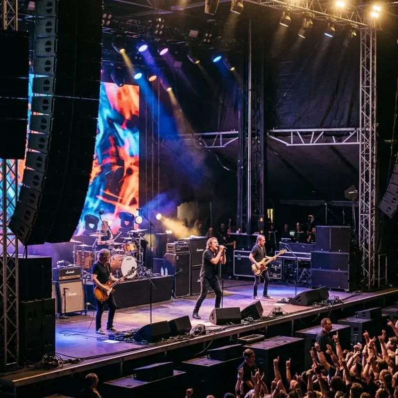Groupe de rock sur scène avec enceintes sono concert en extérieur, public enthousiaste et éclairage coloré.
