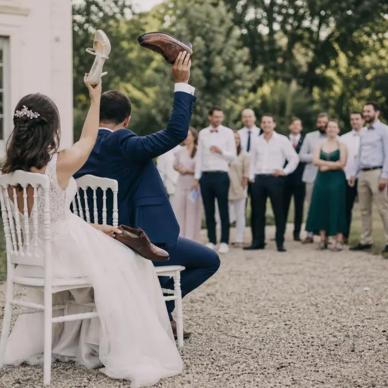 Deux mariés assis sur des chaises, chaussures levées à en l’air, lors d’une animation en plein air devant un groupe d’invités élégamment vêtus. animations originales pour un mariage | scène festive et originale.