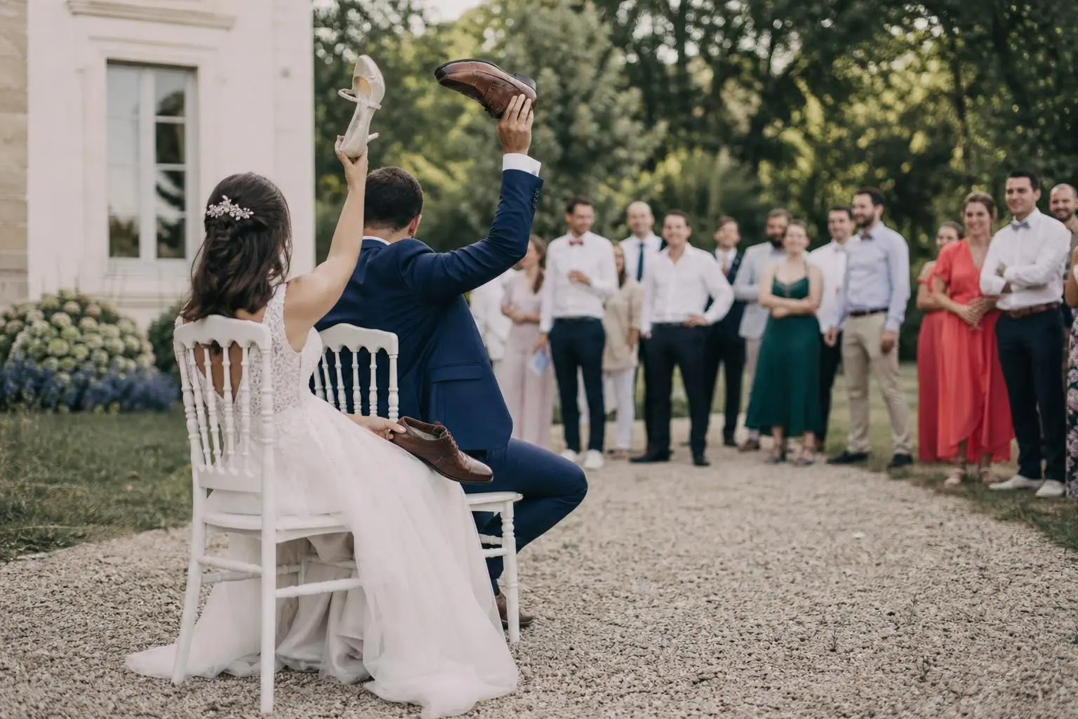 Deux mariés assis sur des chaises, chaussures levées à en l’air, lors d’une animation en plein air devant un groupe d’invités élégamment vêtus. animations originales pour un mariage | scène festive et originale.