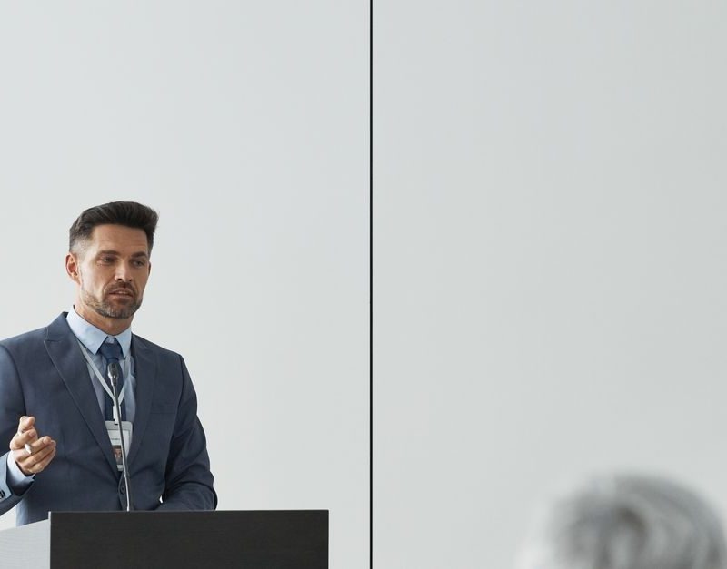 Homme en costume bleu donnant une présentation devant un pupitre lors d’une conférence, public en premier plan.