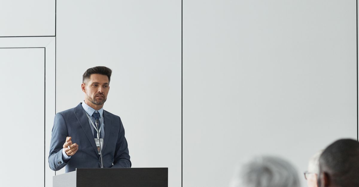 Homme en costume bleu donnant une présentation devant un pupitre lors d’une conférence, public en premier plan.