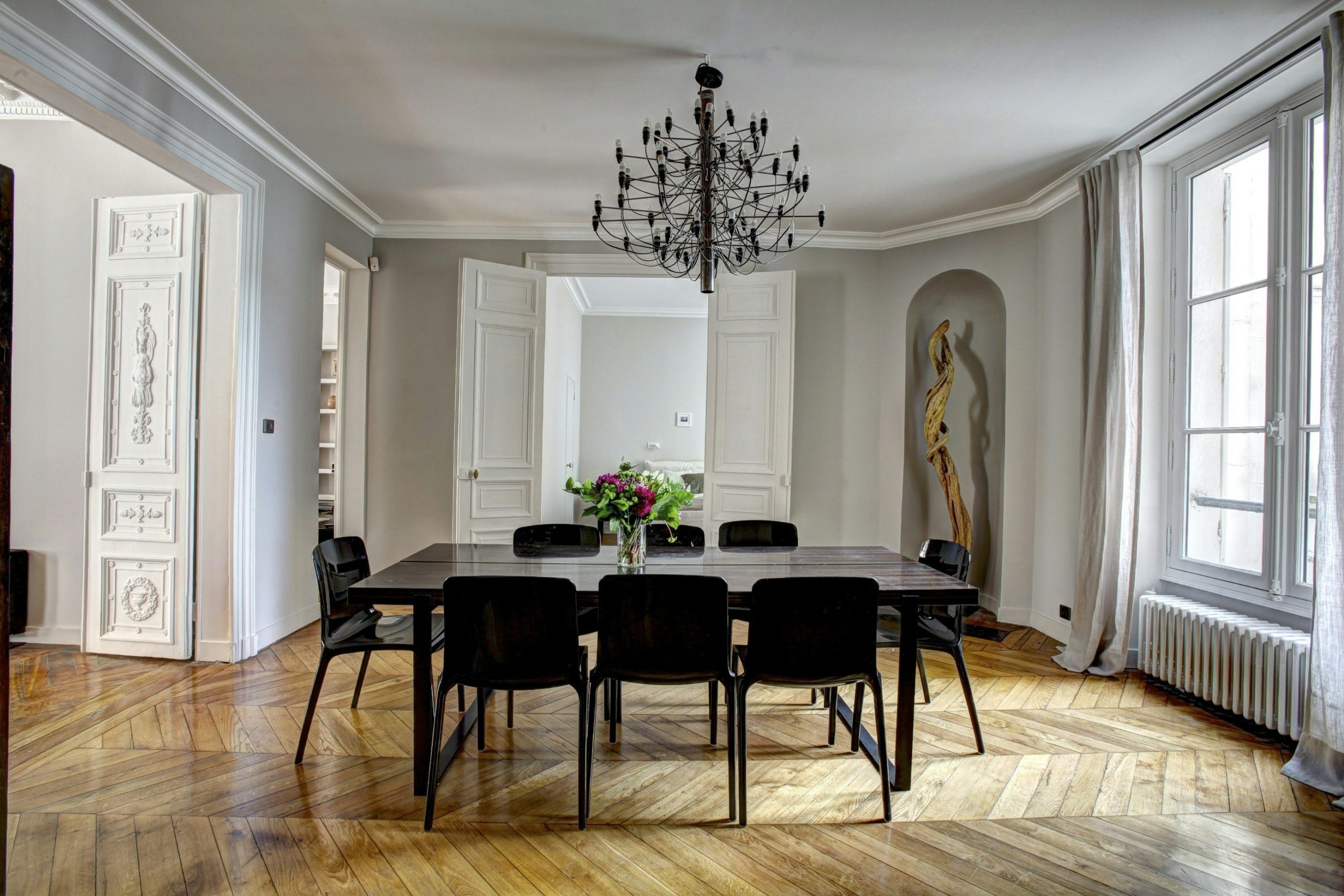 Salle de conférence élégante avec grande table noire et chaises, lustre au plafond et vase de fleurs au centre. Fenêtres lumineuses et parquet en chevrons. Intérieur moderne et lumineux, adapté à un équipement salle de conférence moderne.