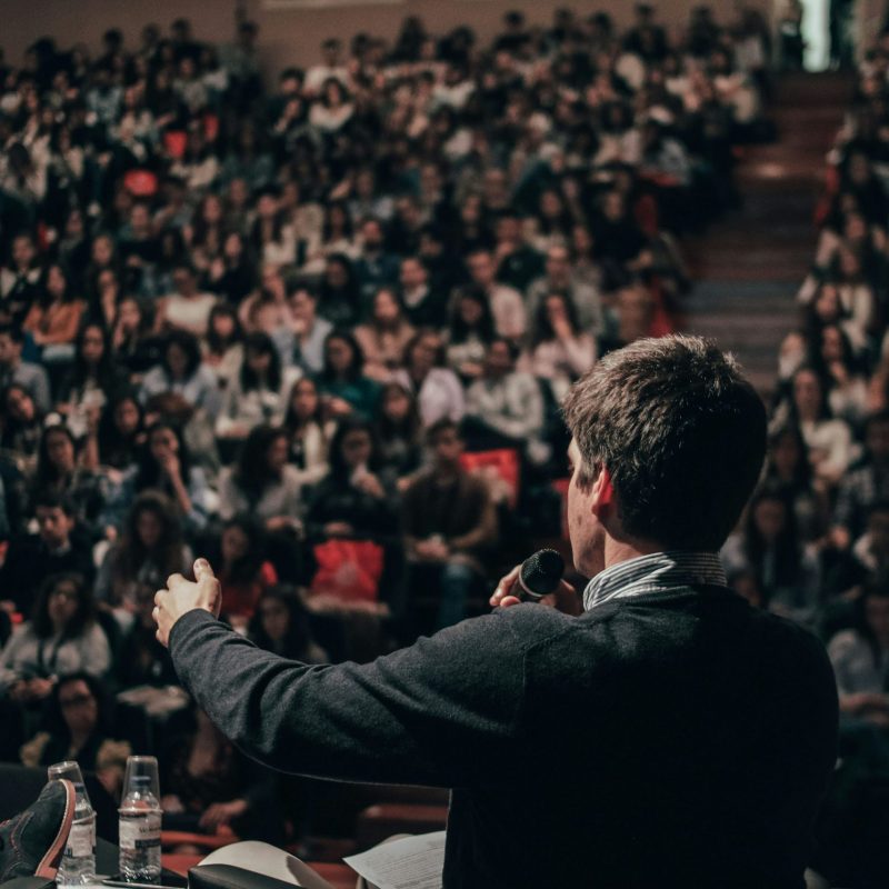 Orateur sur scène, micro à la main, face à une grande foule en salle de conférence, ambiance concentrée et attentive. sonorisation salle de conférence comment choisir