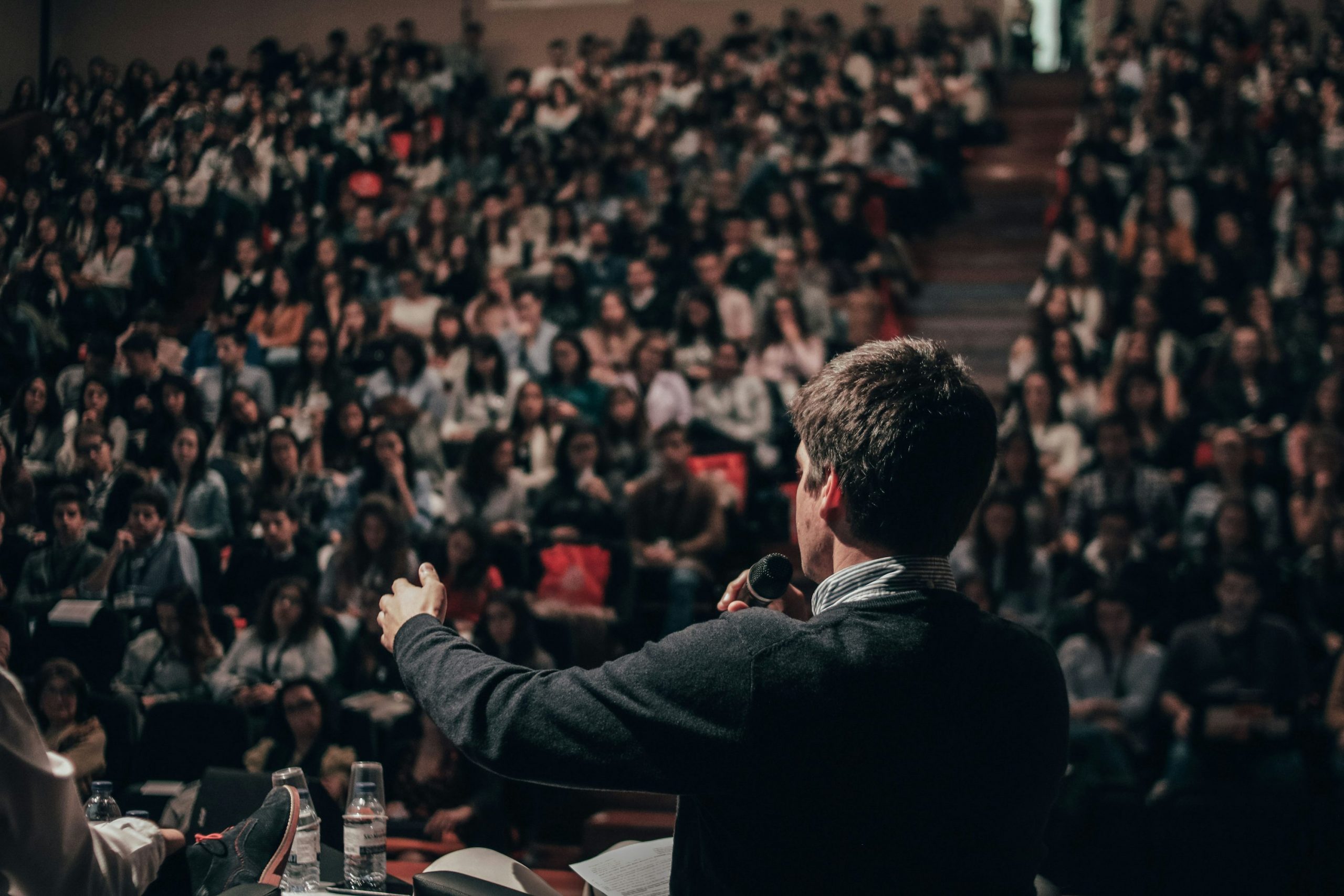 Orateur sur scène, micro à la main, face à une grande foule en salle de conférence, ambiance concentrée et attentive. sonorisation salle de conférence comment choisir