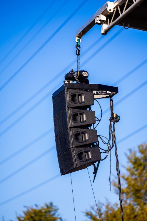 Enceinte de sonorisation suspendue en hauteur, composée de plusieurs caissons, avec câbles et matériel de levage visibles contre un ciel bleu. Puissance sono pour concert en salle.