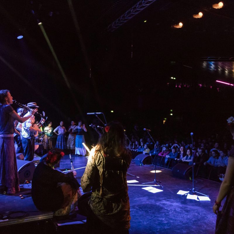 Groupe de musiciens sur scène en salle de concert, public assis et éclairage scénique vibrant. Puissance sono pour concert en salle.