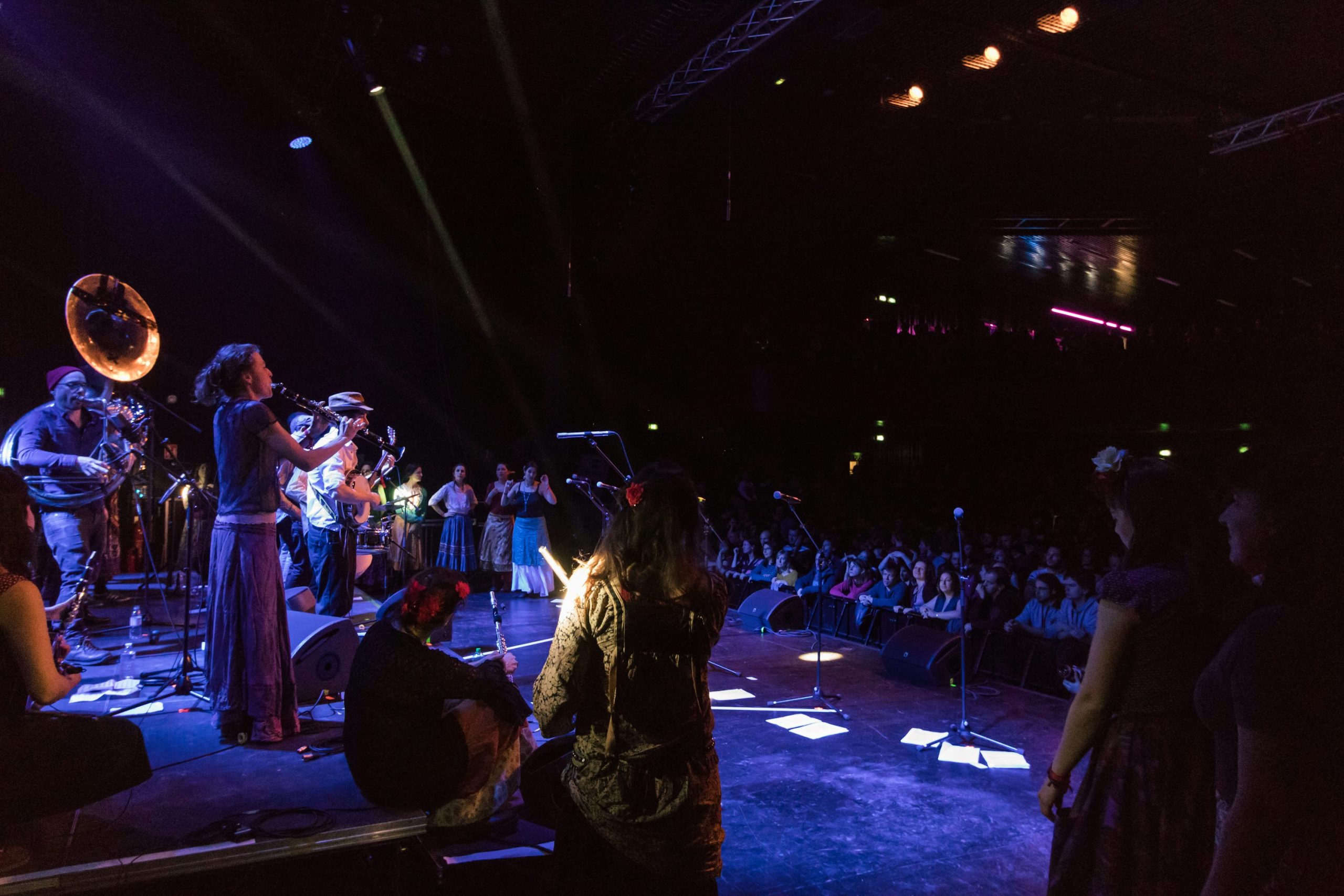 Groupe de musiciens sur scène en salle de concert, public assis et éclairage scénique vibrant. Puissance sono pour concert en salle.