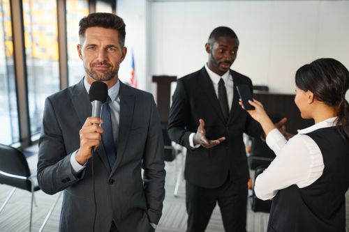 Homme en costume parlant au micro lors d’une conférence politique, deux assistants discutant en arrière-plan. Sonorisation conférence politique dans un cadre professionnel.