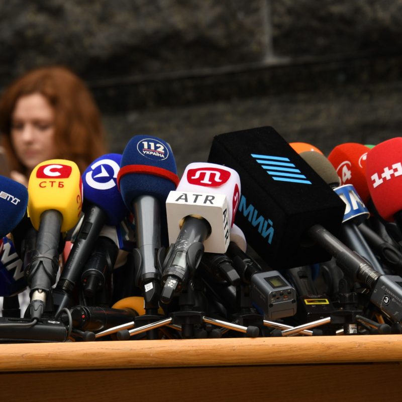 Ras-de-marche de microphones de presse sur un pupitre, avec de nombreux micros colorés de chaînes différentes. Microphone pour conférence non visible mais évoqué par l’ensemble.