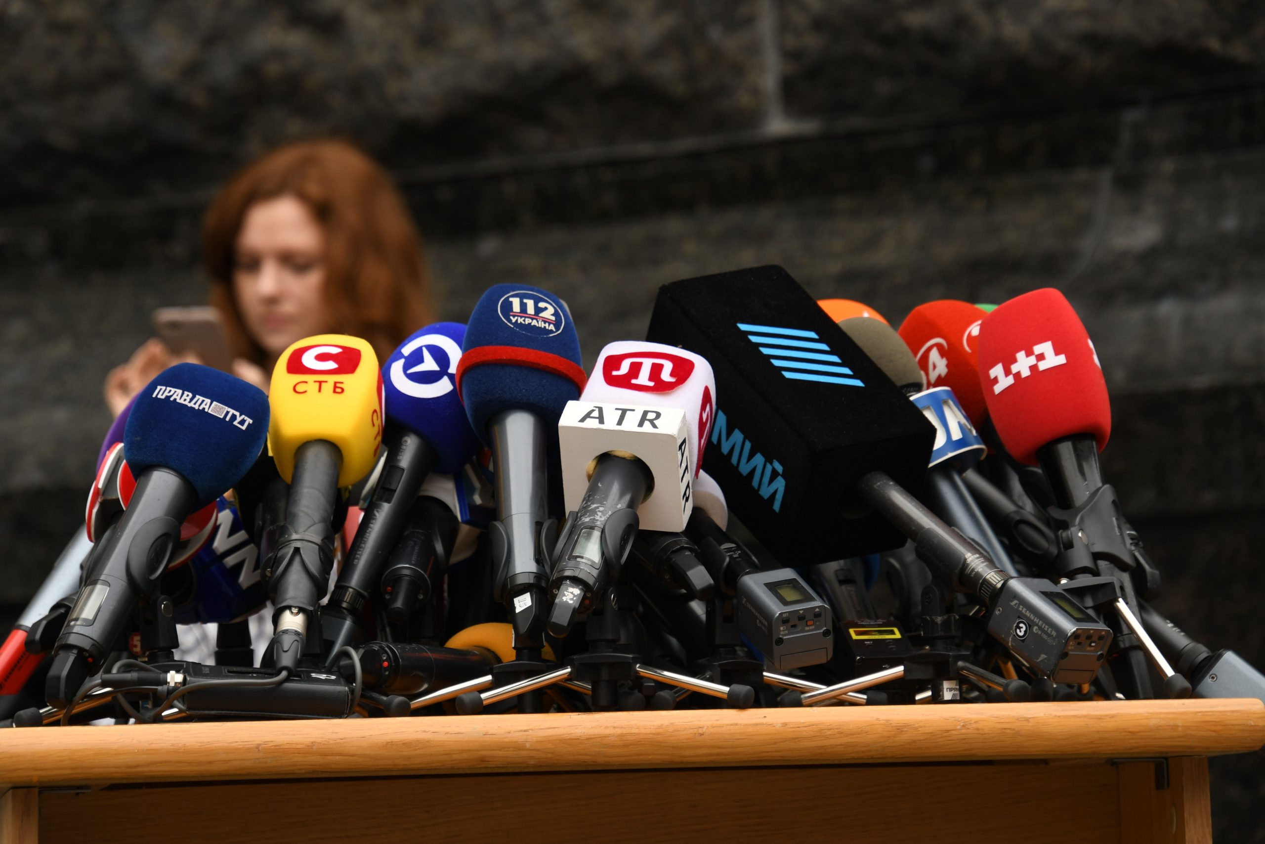 Ras-de-marche de microphones de presse sur un pupitre, avec de nombreux micros colorés de chaînes différentes. Microphone pour conférence non visible mais évoqué par l’ensemble.