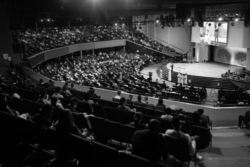 Salle pleine lors d’une conférence politique avec tribune et écran, vue d’ensemble en noir et blanc. Sonorisation conférence politique, public attentif.