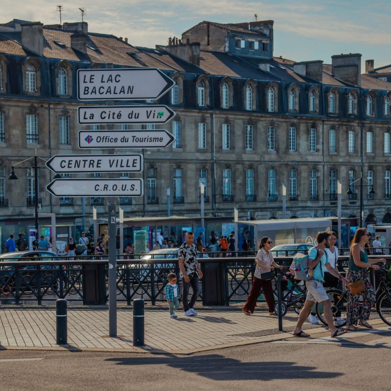 Signage directionnel en front de rive à Bordeaux, passant·e·s et vélos près d’un pont, façades anciennes en arrière-plan. agence événementielle bordeaux gironde intégrée.