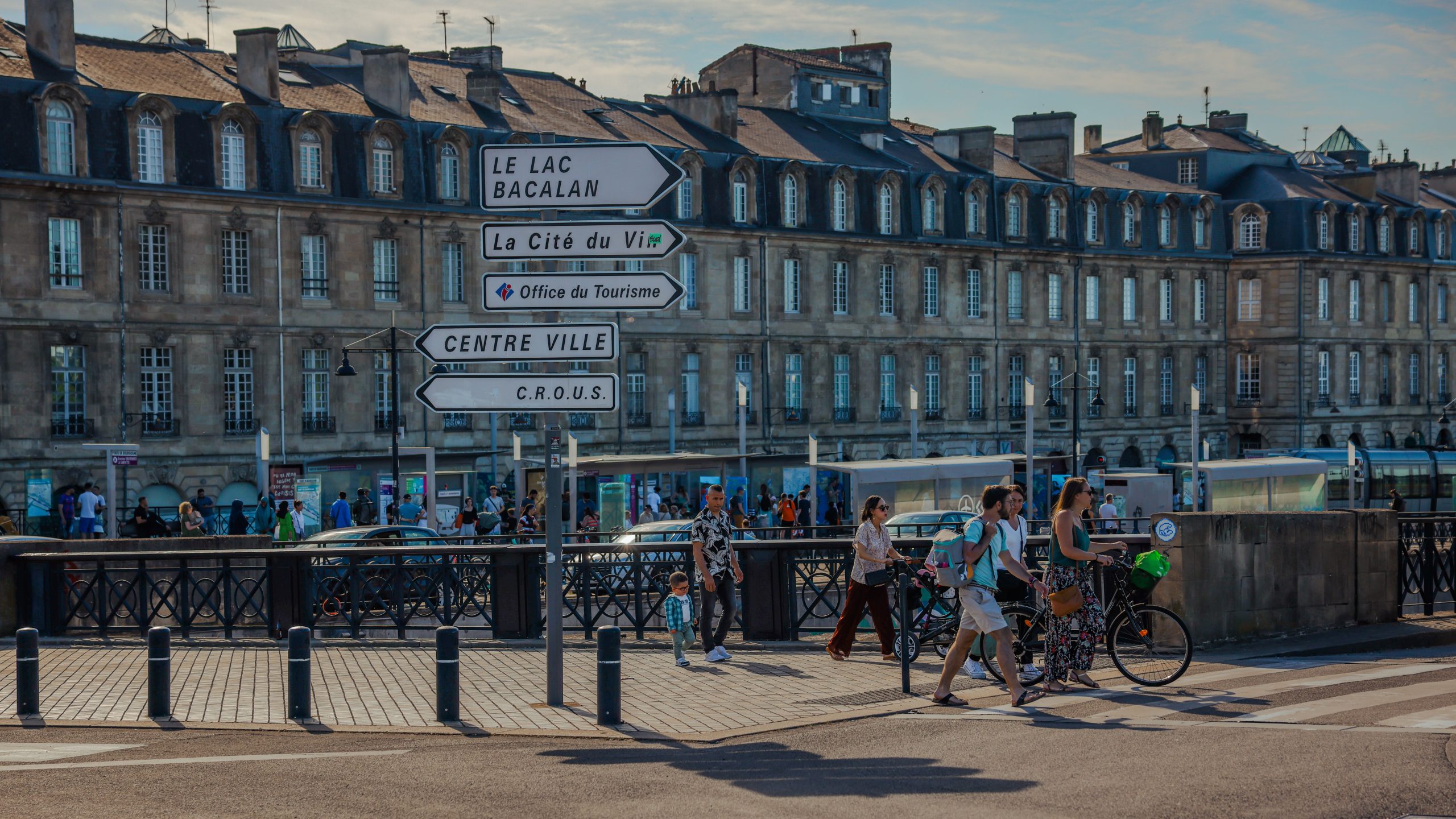 Signage directionnel en front de rive à Bordeaux, passant·e·s et vélos près d’un pont, façades anciennes en arrière-plan. agence événementielle bordeaux gironde intégrée.