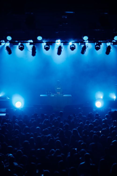 Spectacle en intérieur avec une scène éclairée en bleu et une foule dense, ambiance club, possible DJ en arrière-plan. Conception éclairage événement professionnel évoquée.