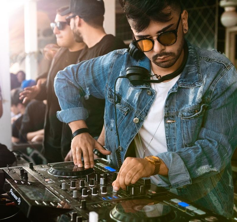 DJ professionnel en train de mixer sur une platine avec casque autour du cou et lunettes de soleil.