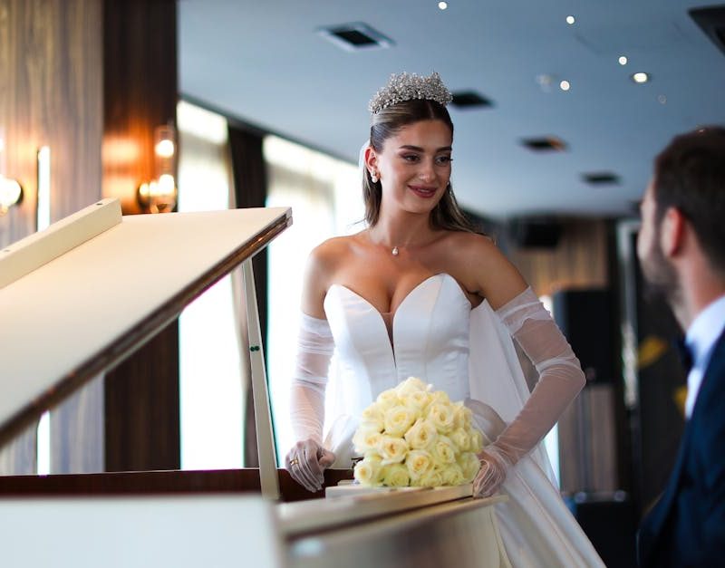 Mariée en robe blanche et diadème, debout au piano à queue, tenant bouquet de roses blanches et échangeant un sourire avec