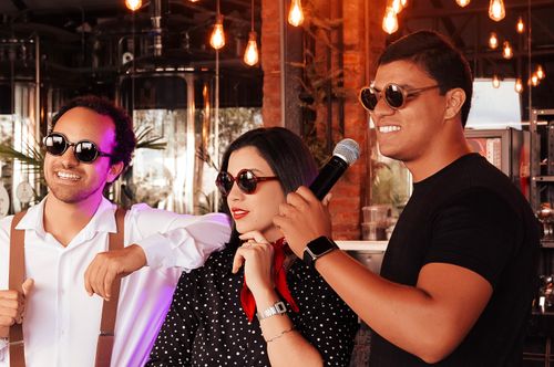 Trois chanteurs portant des lunettes de soleil sourient, l’un tient un micro près de sa bouche.